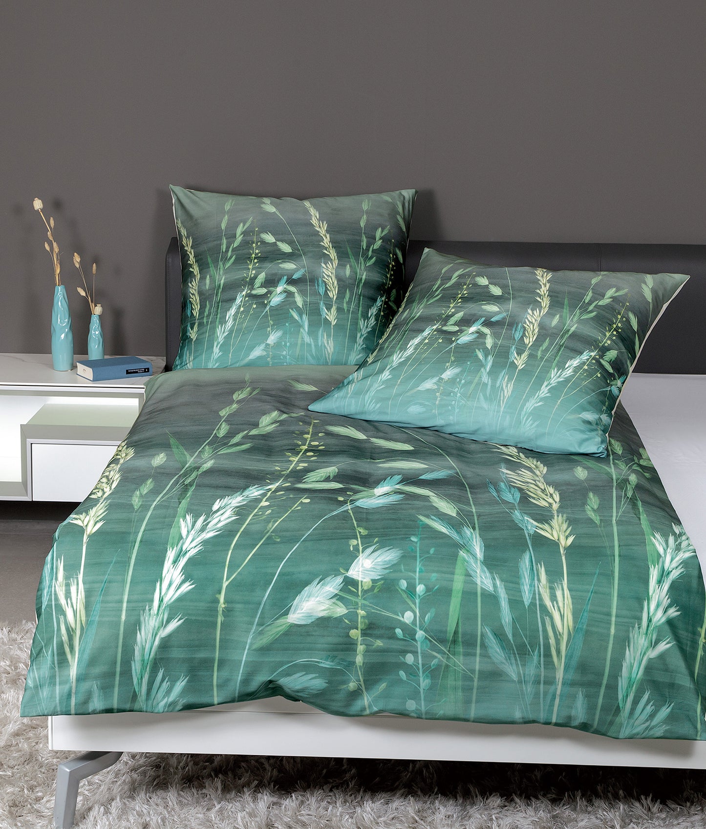 Auf einem Bett liegen mehrere Kopfkissen und eine Bettdecke bezogen mit Mako-Satin Bettwaesche aus 100 Prozent Baumwolle mit Muster aus Graesern in der Farbe Gruen. Modell Modern Art Green.