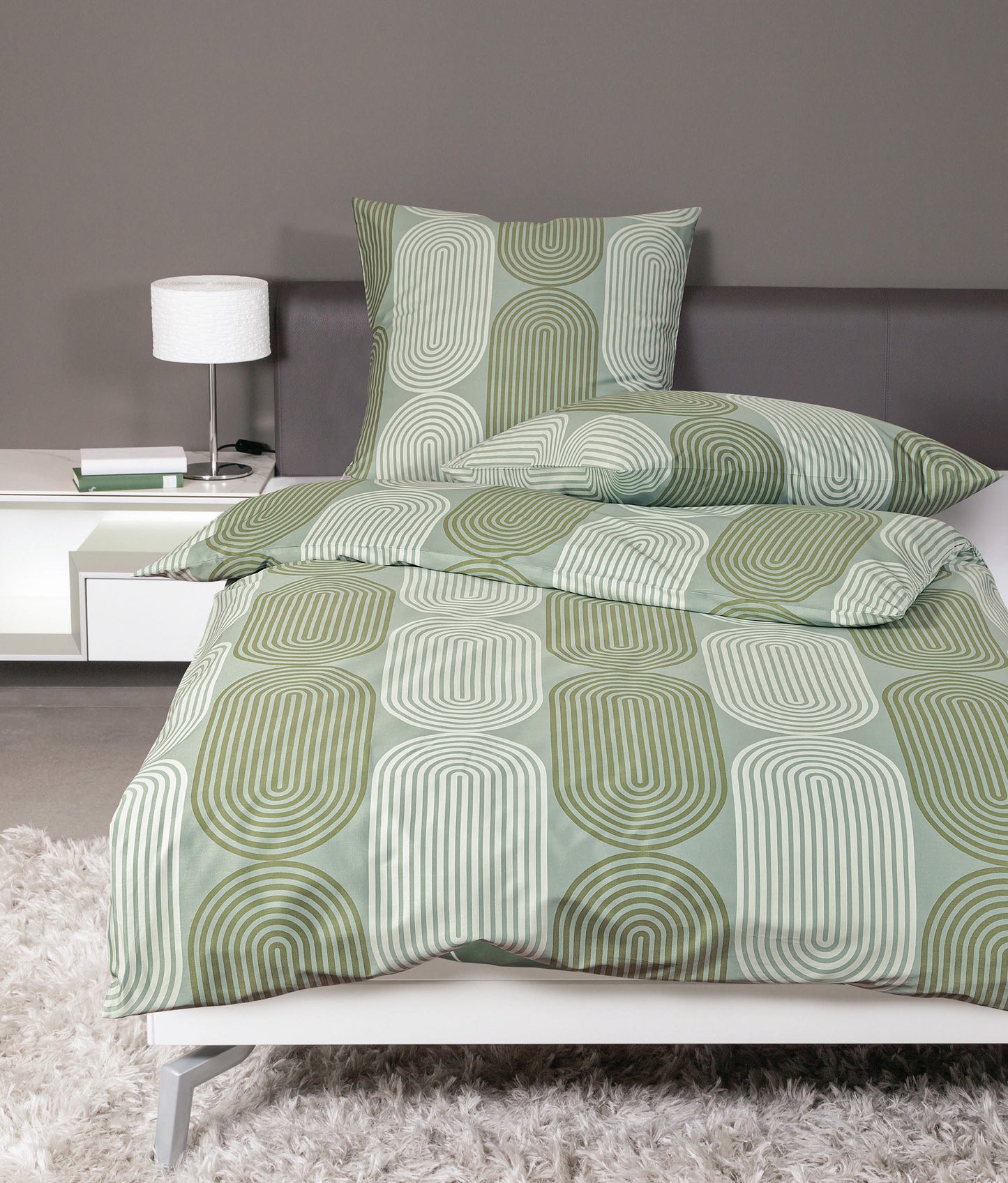 Auf einem Bett liegen mehrere Kopfkissen und eine Bettdecke bezogen mit Mako-Satin Bettwaesche aus 100 Prozent Baumwolle mit Muster aus grafischen Formen in der Farbe Gruen. Modell Rauchgrün Lind.