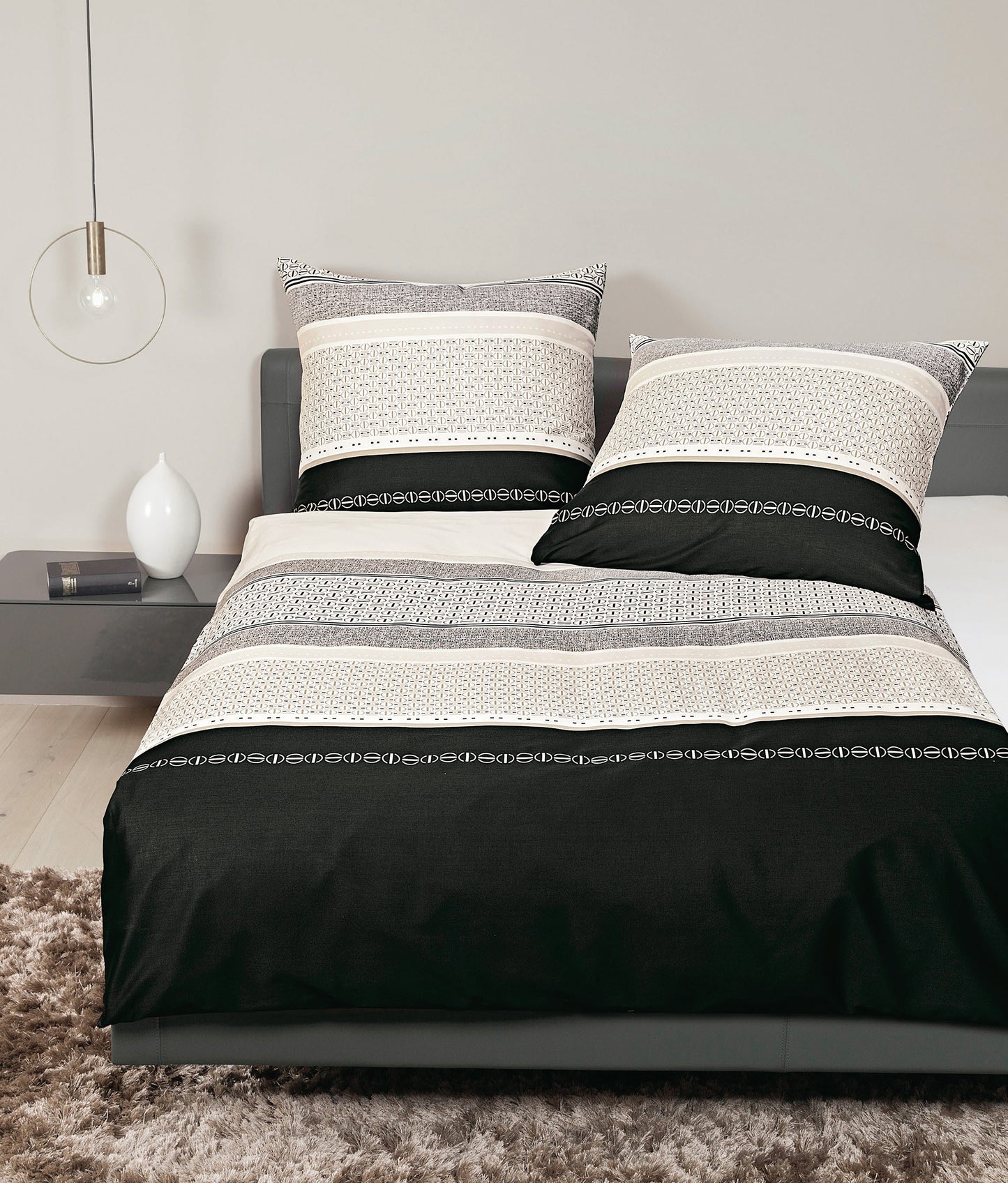 Auf einem Bett liegen mehrere Kopfkissen und eine Bettdecke bezogen mit Mako-Satin Bettwaesche aus 100 Prozent Baumwolle mit Muster aus grafischen Formen in der Farbe Schwarz. Modell Schwarz Kitt.