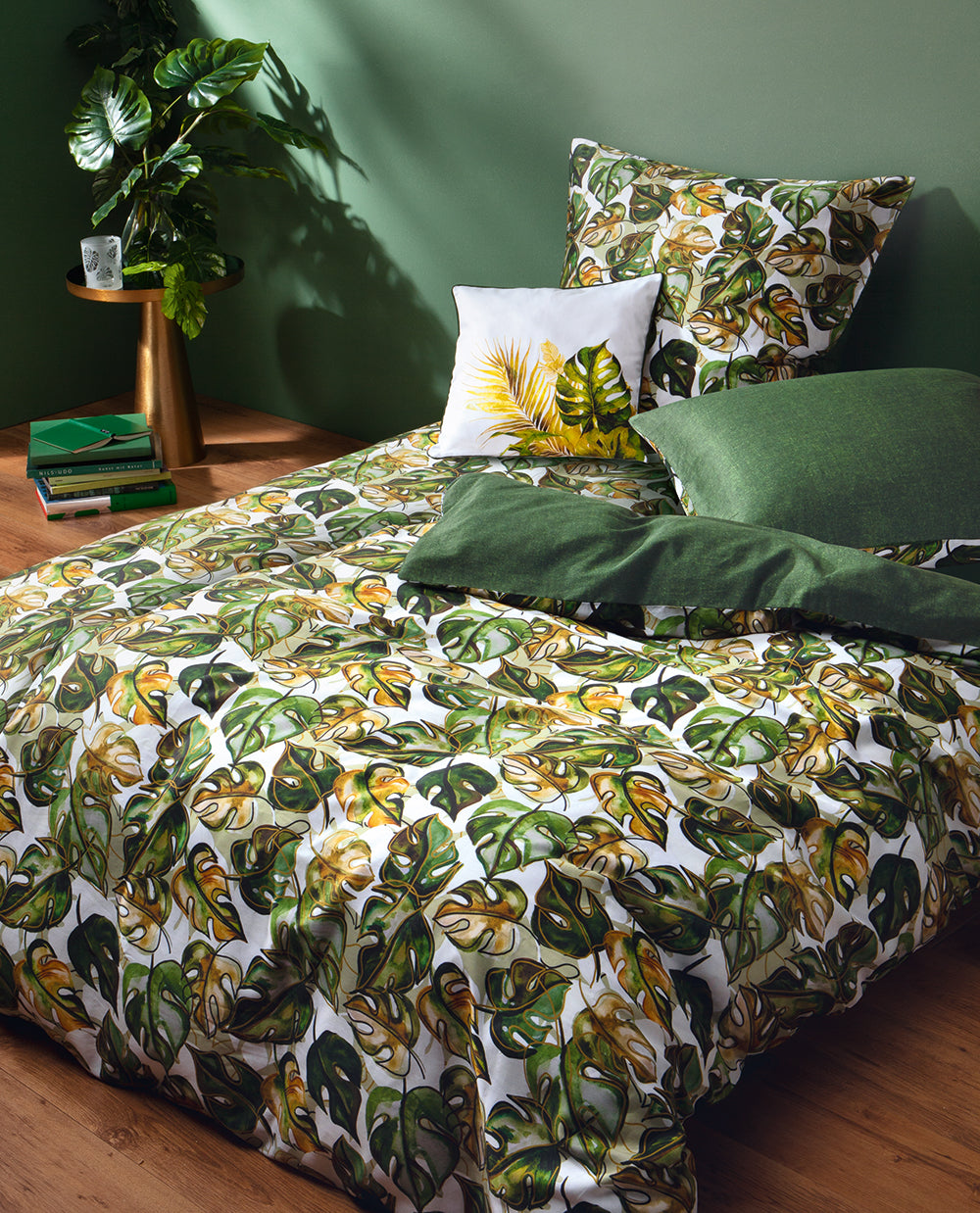 Auf einem Bett liegen mehrere Kopfkissen und eine Bettdecke bezogen mit Satin Bettwaesche aus 100 Prozent Baumwolle mit Muster aus Blaettern in der Farbe Gruen. Modell Verdea.