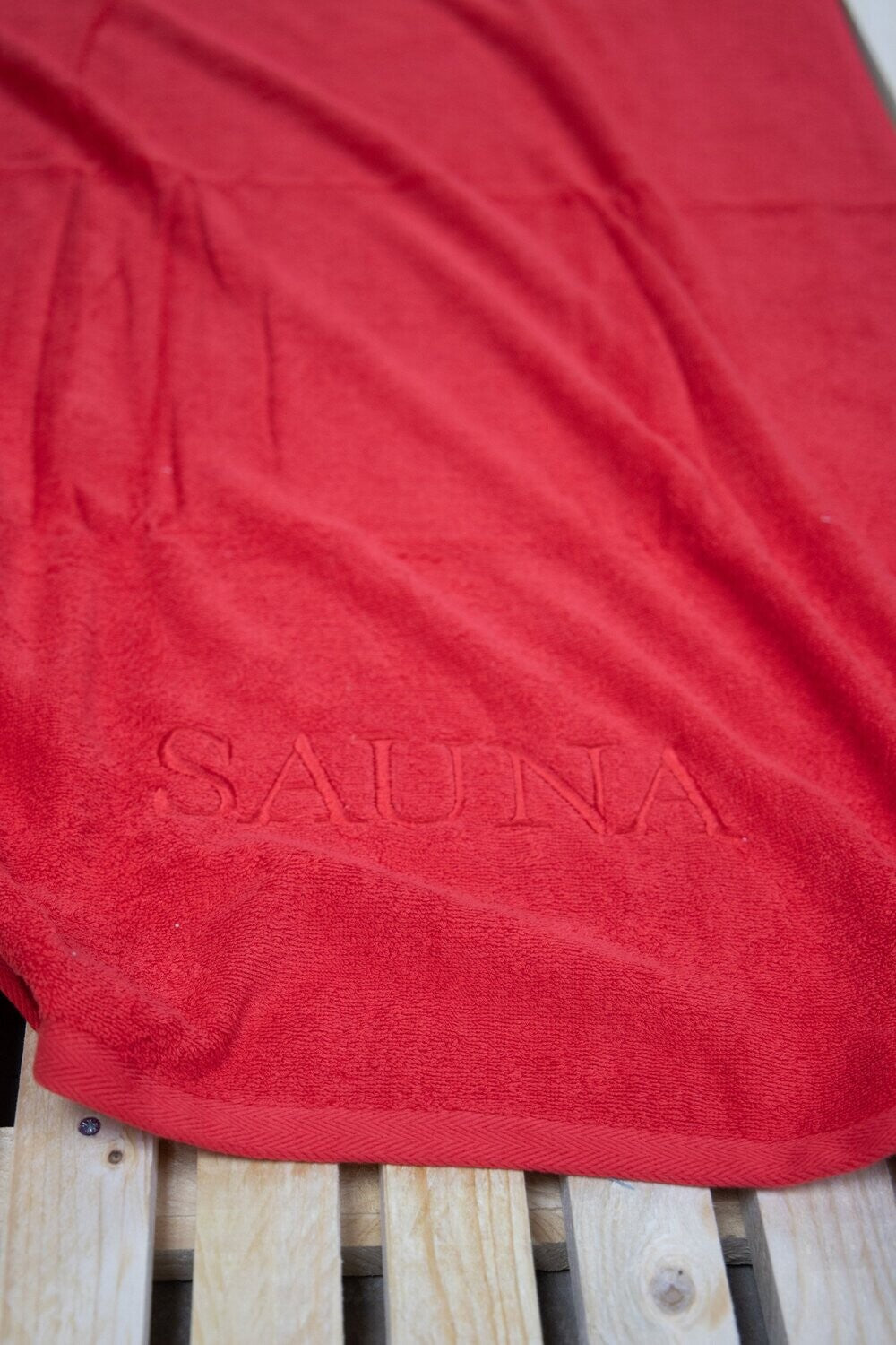 Auf einer Bank liegt ein Saunatuch mit aufgesticktem Text SAUNA von p.k. aus 100 Prozent Baumwolle in der Groesse 70x200cm. Farbe Rot. Modell Rot.