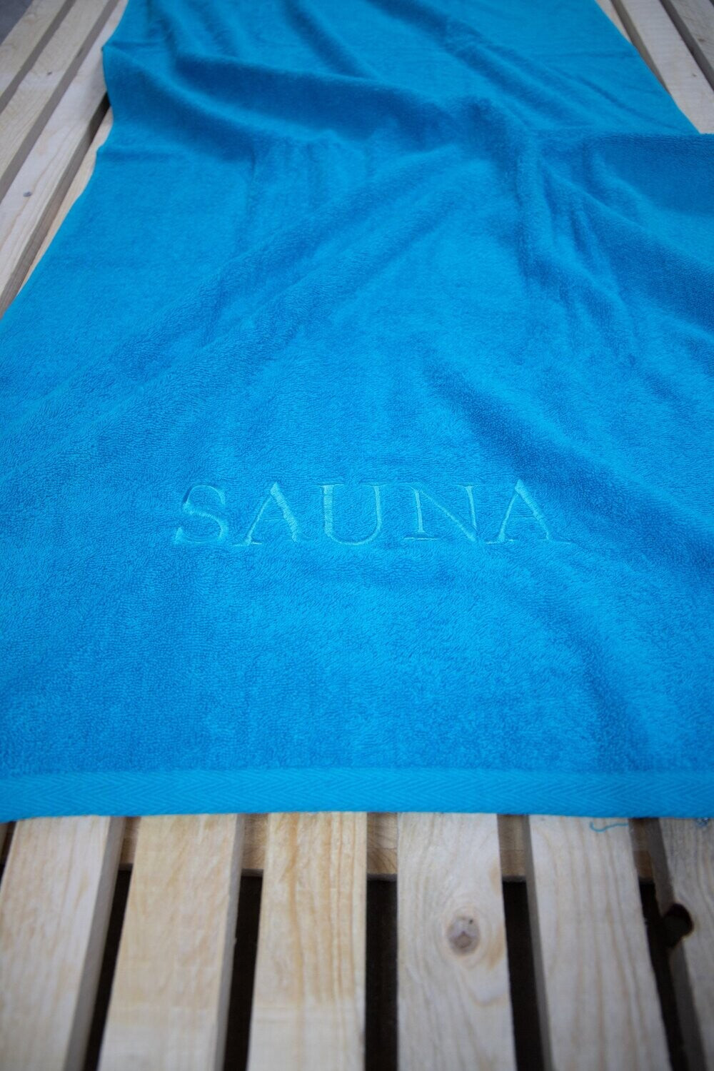 Auf einer Bank liegt ein Saunatuch mit aufgesticktem Text SAUNA von p.k. aus 100 Prozent Baumwolle in der Groesse 70x200cm. Farbe Tuerkis. Modell Tuerkis.