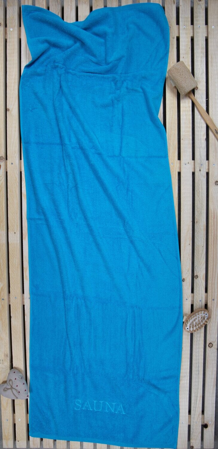 Auf einer Holzbankank liegt ein Saunatuch mit aufgesticktem Text SAUNA von p.k. aus 100 Prozent Baumwolle in der Groesse 70x200cm. Farbe Tuerkis. Modell Tuerkis.