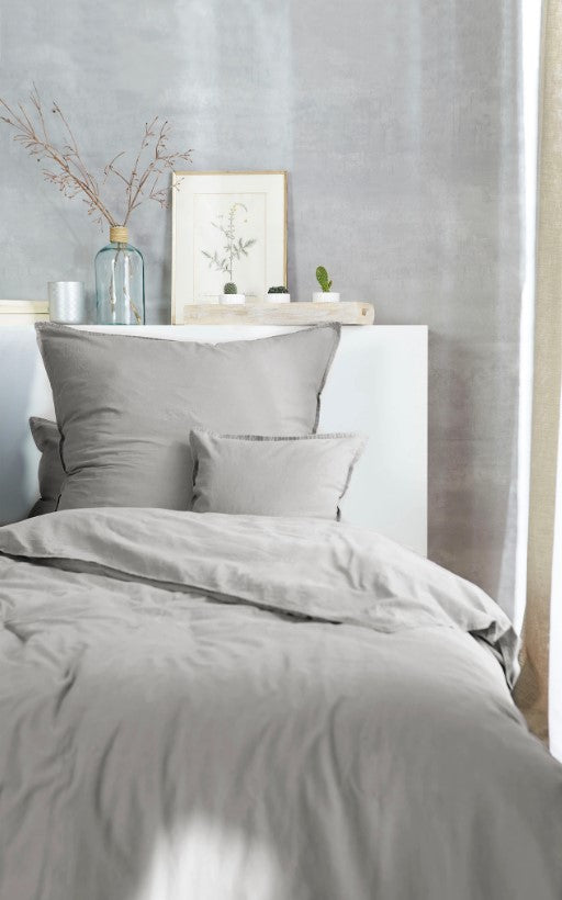 Auf einem Bett liegen mehrere Kopfkissen und eine Bettdecke bezogen mit Renforce Bettwaesche aus 100 Prozent Baumwolle in der Farbe Silber. Modell Stone-Washed Hellgrau 722.