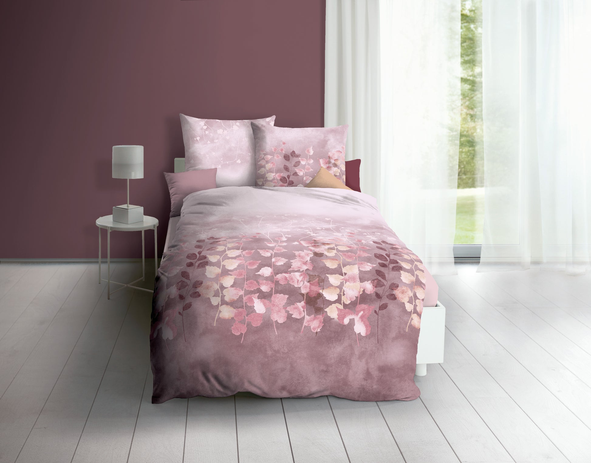 Auf einem Bett liegen mehrere Kopfkissen und eine Bettdecke bezogen mit Biber Bettwaesche aus 100 Prozent Baumwolle mit Muster aus Pflanzen in der Farbe Rosa. Modell Mauve.