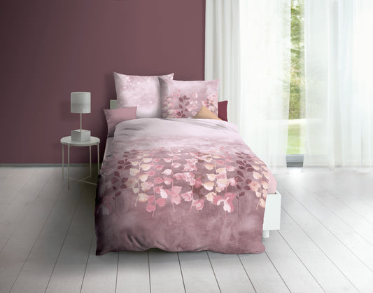 Auf einem Bett liegen mehrere Kopfkissen und eine Bettdecke bezogen mit Biber Bettwaesche aus 100 Prozent Baumwolle mit Muster aus Pflanzen in der Farbe Rosa. Modell Mauve.