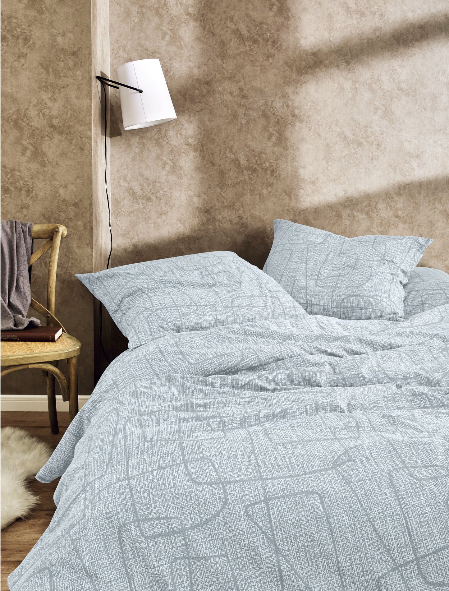 Auf einem Bett liegen mehrere Kopfkissen und eine Bettdecke bezogen mit Edel-Feinbiber Bettwaesche aus 100 Prozent Baumwolle mit Muster aus Linien in der Farbe Grau. Modell Geometrisch.