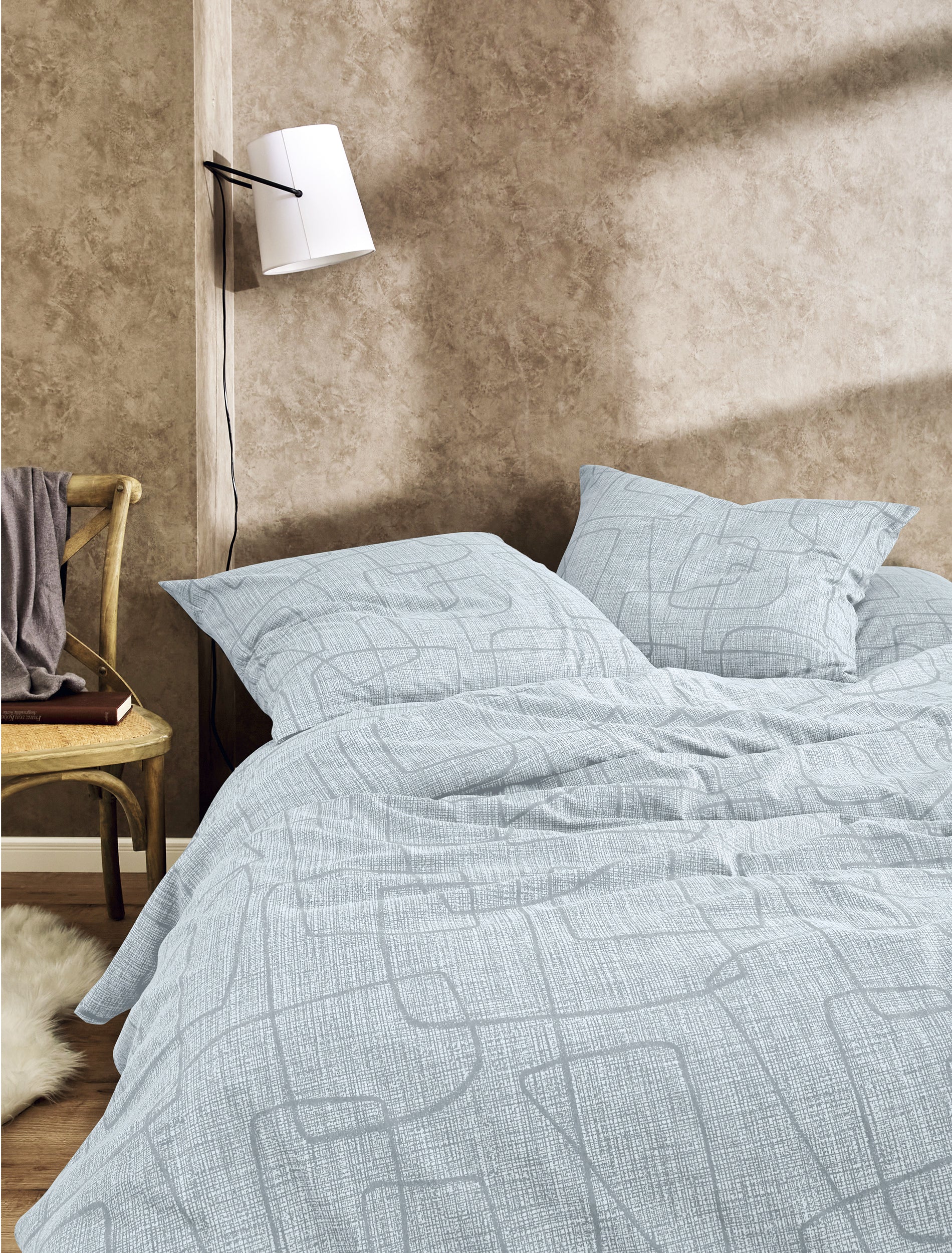 Auf einem Bett liegen mehrere Kopfkissen und eine Bettdecke bezogen mit Edel-Feinbiber Bettwaesche aus 100 Prozent Baumwolle mit Muster aus Linien in der Farbe Grau. Modell Geometrisch.