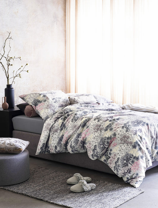 Auf einem Bett liegen mehrere Kopfkissen und eine Bettdecke bezogen mit Edel-Feinbiber Bettwaesche aus 100 Prozent Baumwolle mit Muster aus Blumen in der Farbe Silber. Modell Flowers.
