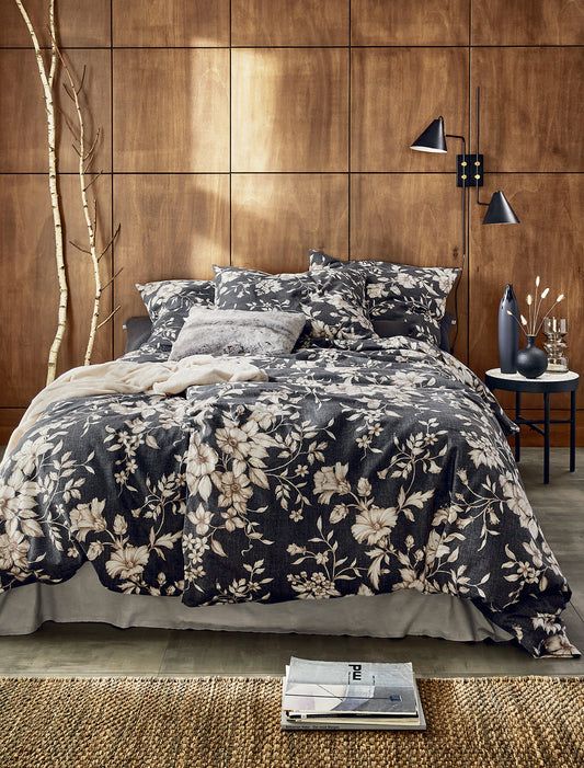 Auf einem Bett liegen mehrere Kopfkissen und eine Bettdecke bezogen mit Edel-Feinbiber Bettwaesche aus 100 Prozent Baumwolle mit Muster aus Blumen in der Farbe Grau. Modell Grau.