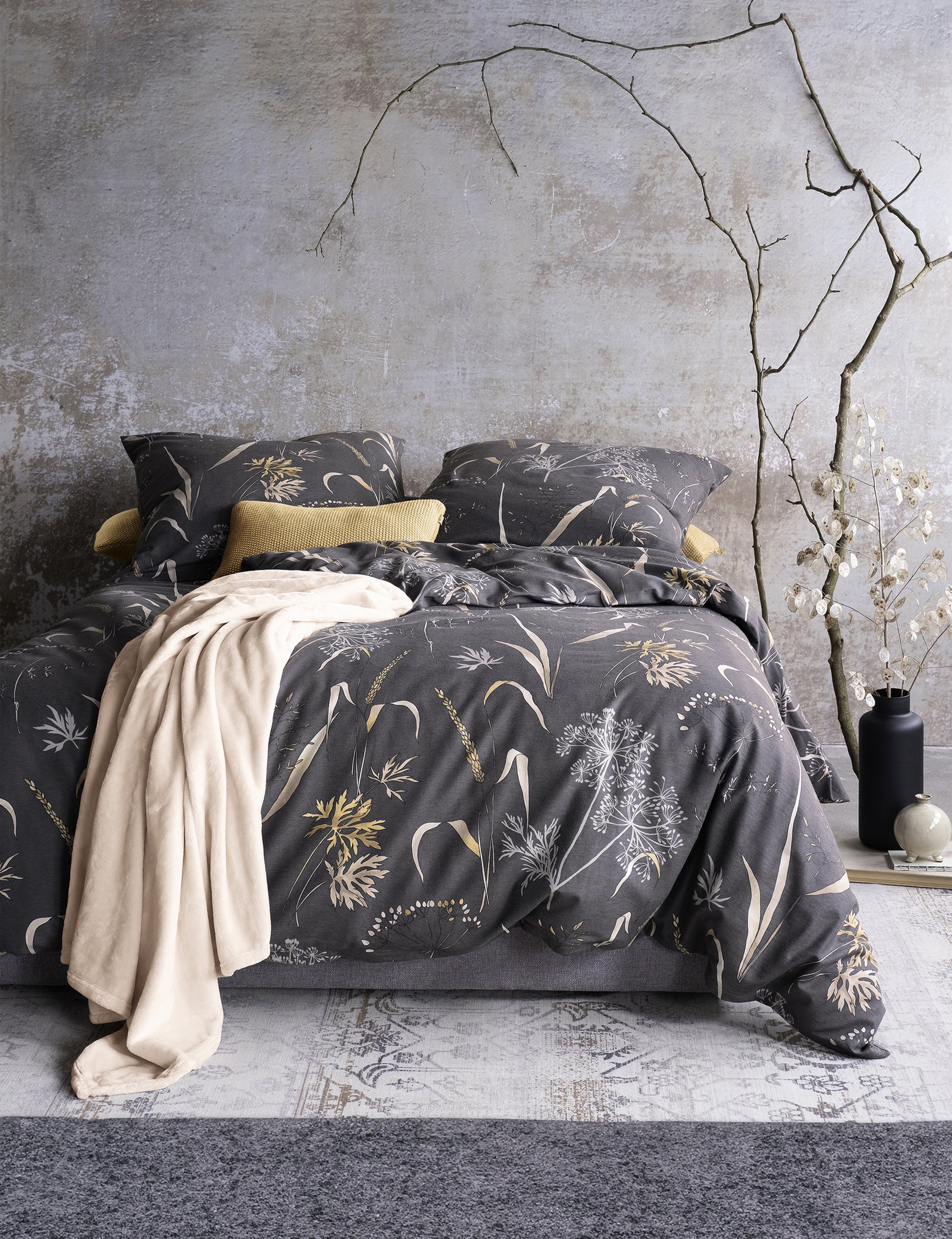 Auf einem Bett liegen mehrere Kopfkissen und eine Bettdecke bezogen mit Edel-Feinbiber Bettwaesche aus 100 Prozent Baumwolle mit Muster aus Graesern in der Farbe Grau. Modell Luzara.