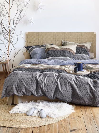 Auf einem Bett liegen mehrere Kopfkissen und eine Bettdecke bezogen mit Edel-Feinbiber Bettwaesche aus 100 Prozent Baumwolle mit Muster aus geographischen Formen in der Farbe Blau. Modell Panda Blau.