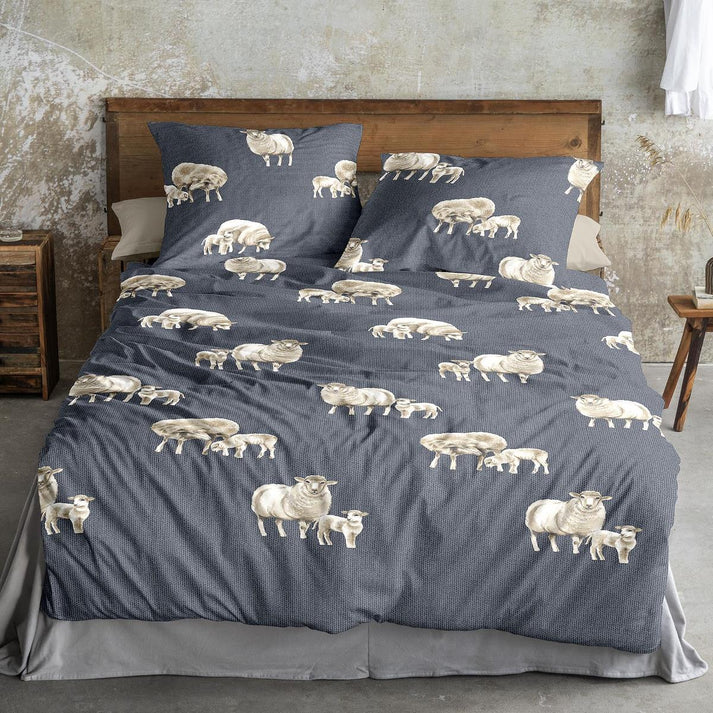 Auf einem Bett liegen mehrere Kopfkissen und eine Bettdecke bezogen mit Edel-Feinbiber Bettwaesche aus 100 Prozent Baumwolle mit Muster aus SChaefchen in der Farbe Blau. Modell Schaefchen Blau.