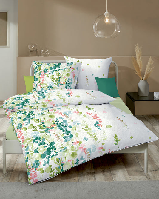 Auf einem Bett liegen mehrere Kopfkissen und eine Bettdecke bezogen mit Edel Seersucker Bettwaesche aus 100 Prozent Baumwolle mit Muster aus Pflanzen in der Farbe Weiss. Modell 466/403 Gruen.