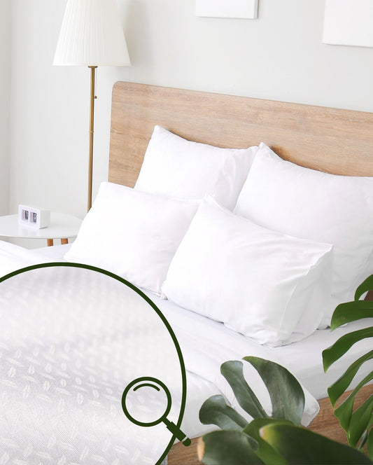 Ein Bett mit weisser Bettwaesche mit Strukturmuster und vier Kissen, umgeben von einer Pflanze, einer Lampe und einem Wecker.