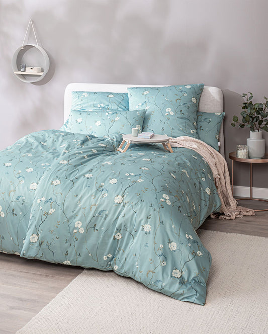 Ein Bettbezug-Set aus mehreren Kopfkissen und einer Bettdecke bezogen mit Mako-Satin Bettwaesche aus 100 Prozent Baumwolle mit Muster aus Blumen in der Farbe Petrol. Modell Soraya Petrol.