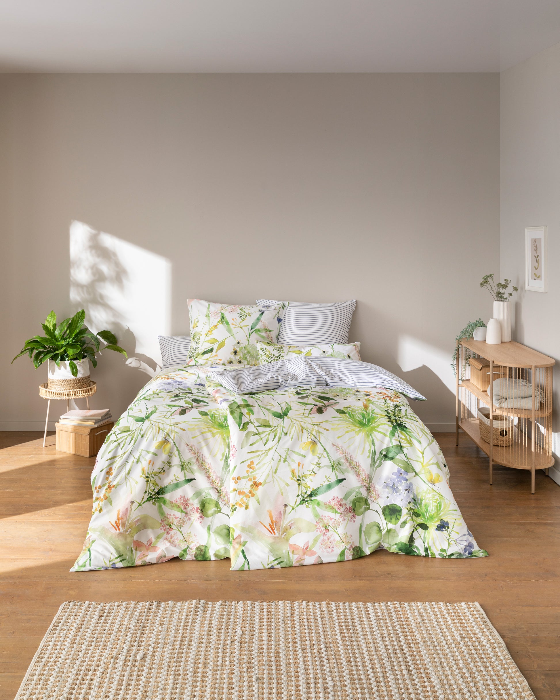 Auf einem Bett liegen mehrere Kopfkissen und eine Bettdecke bezogen mit Mako-Satin Bettwaesche aus 100 Prozent Baumwolle mit Muster aus Blumen in der Farbe Weiss. Modell Summer.