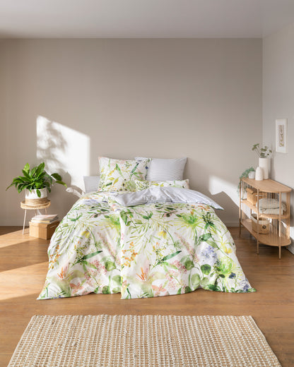Auf einem Bett liegen mehrere Kopfkissen und eine Bettdecke bezogen mit Mako-Satin Bettwaesche aus 100 Prozent Baumwolle mit Muster aus Blumen in der Farbe Weiss. Modell Summer.