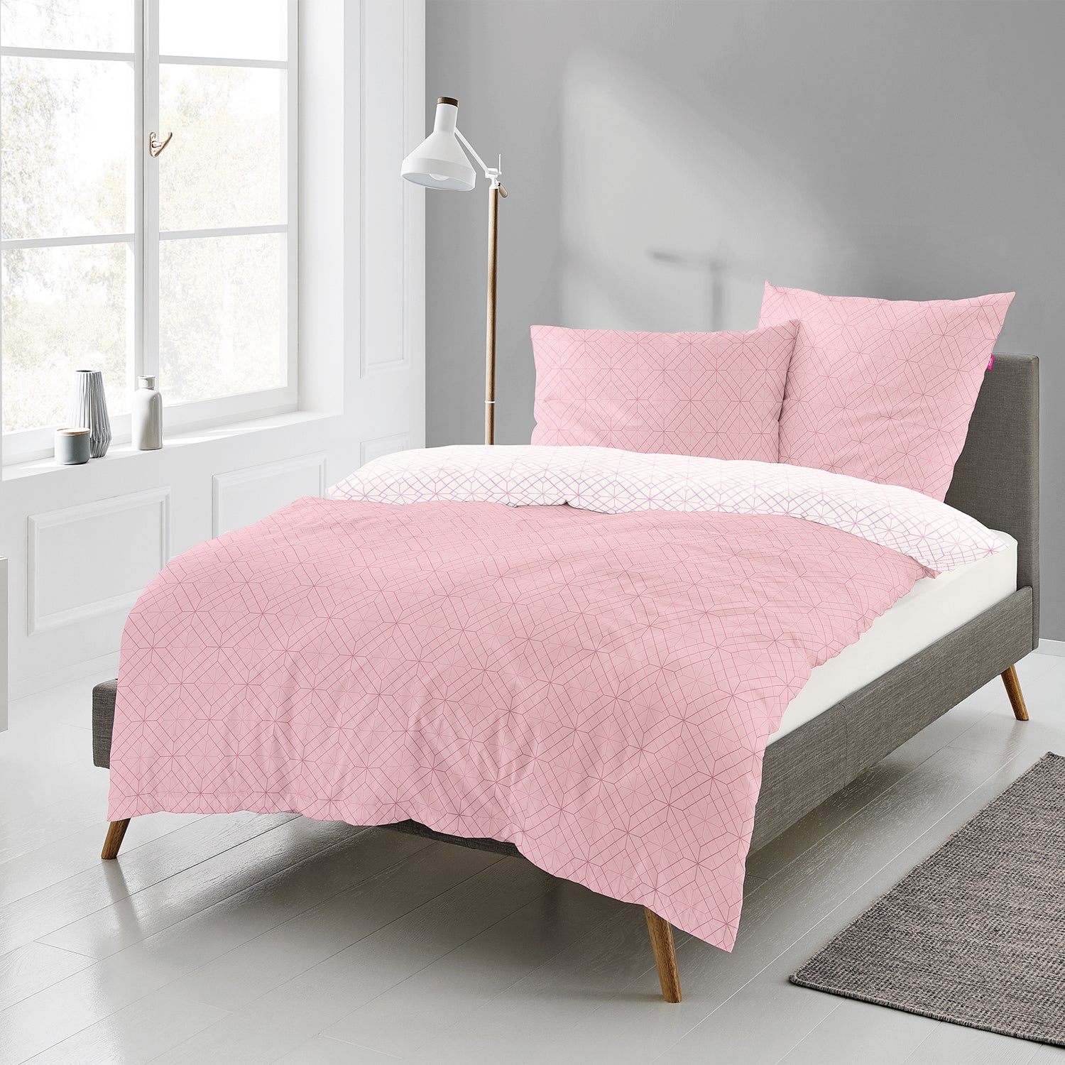 Auf einem Bett liegen mehrere Kopfkissen und eine Bettdecke bezogen mit Mako-Satin Bettwaesche aus 100 Prozent Baumwolle mit Muster aus geografischen Formen in der Farbe Rosa. Modell Freundin Home Collection Corado.