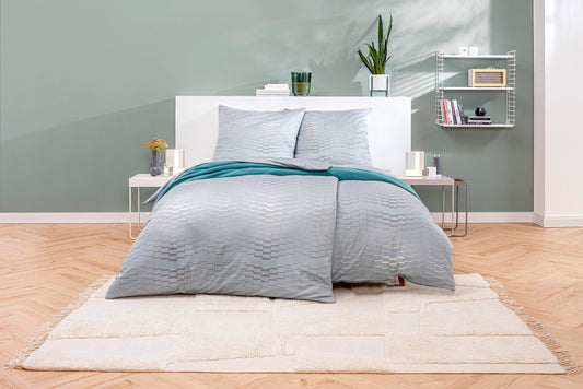 Ein Bettbezug-Set aus mehreren Kopfkissen und einer Bettdecke bezogen mit Mako-Interlock-Jersey Bettwaesche aus 100 Prozent Baumwolle mit Muster aus horizontalen Zick-Zack Linien in der Farbe Silber. Modell Marian.