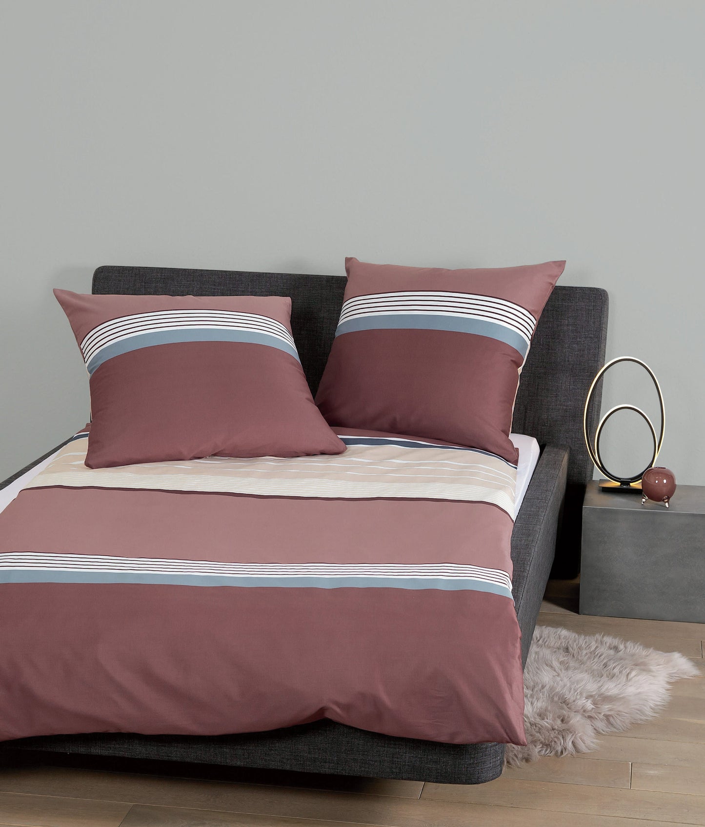 Auf einem Bett liegen mehrere Kopfkissen und eine Bettdecke bezogen mit Mako-Satin Bettwaesche aus 100 Prozent Baumwolle mit Muster aus horizontalen Streifen in der Farbe Bordeaux. Modell Burgund Rosé.