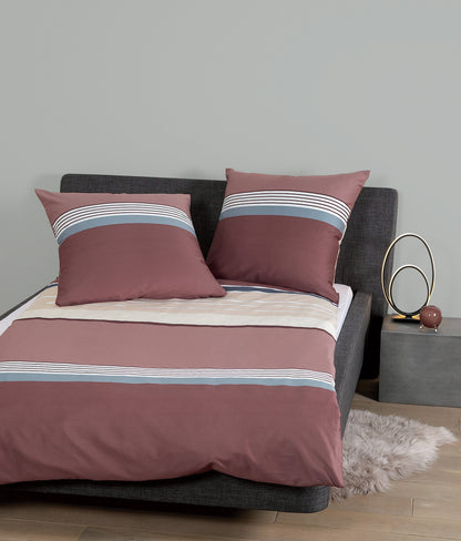 Auf einem Bett liegen mehrere Kopfkissen und eine Bettdecke bezogen mit Mako-Satin Bettwaesche aus 100 Prozent Baumwolle mit Muster aus horizontalen Streifen in der Farbe Bordeaux. Modell Burgund Rosé.