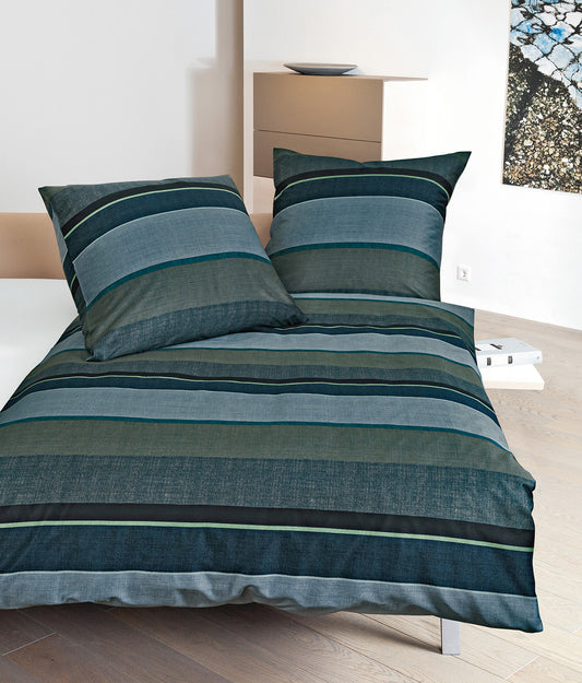 Auf einem Bett liegen mehrere Kopfkissen und eine Bettdecke bezogen mit Mako-Satin Bettwaesche aus 100 Prozent Baumwolle mit Muster aus horizontalen Streifen in der Farbe Blau. Modell Moments Blue.