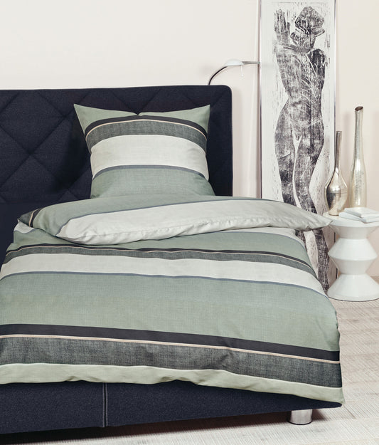 Auf einem Bett liegt ein Kopfkissen und eine Bettdecke bezogen mit Mako-Satin Bettwaesche aus 100 Prozent Baumwolle mit Muster aus horizontalen Streifen in der Farbe Gruen. Modell Moments Mondnebel Jadehauch.
