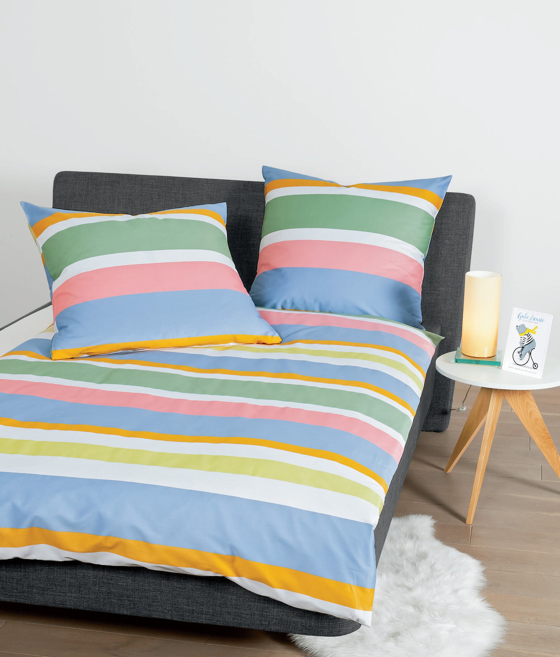 Auf einem Bett liegen mehrere Kopfkissen und eine Bettdecke bezogen mit Mako-Satin Bettwaesche aus 100 Prozent Baumwolle mit Muster aus horizontalen Streifen in der Farbe Mehrfarbig. Modell Multicolor.