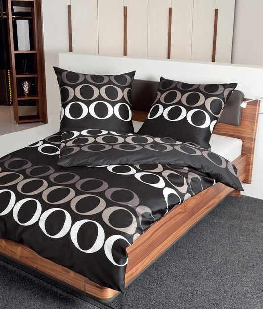 Auf einem Bett liegen mehrere Kopfkissen und eine Bettdecke bezogen mit Mako-Satin Bettwaesche aus 100 Prozent Baumwolle mit Muster aus grafischen Formen in der Farbe Schwarz. Modell Palermo Orbita.
