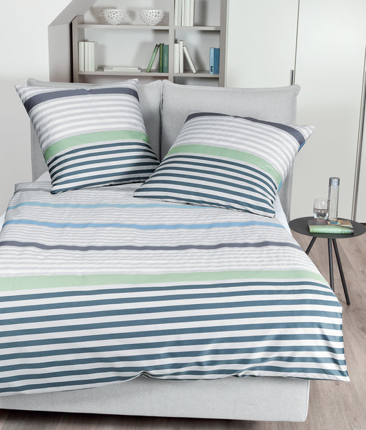 Auf einem Bett liegen mehrere Kopfkissen und eine Bettdecke bezogen mit Mako-Satin Bettwaesche aus 100 Prozent Baumwolle mit Muster aus horizontalen Streifen in der Farbe Mehrfarbig. Modell Perlblau Jade.