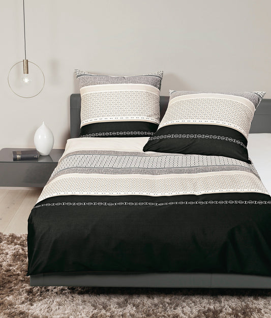 Auf einem Bett liegen mehrere Kopfkissen und eine Bettdecke bezogen mit Mako-Satin Bettwaesche aus 100 Prozent Baumwolle mit Muster aus grafischen Formen in der Farbe Schwarz. Modell Schwarz Kitt.