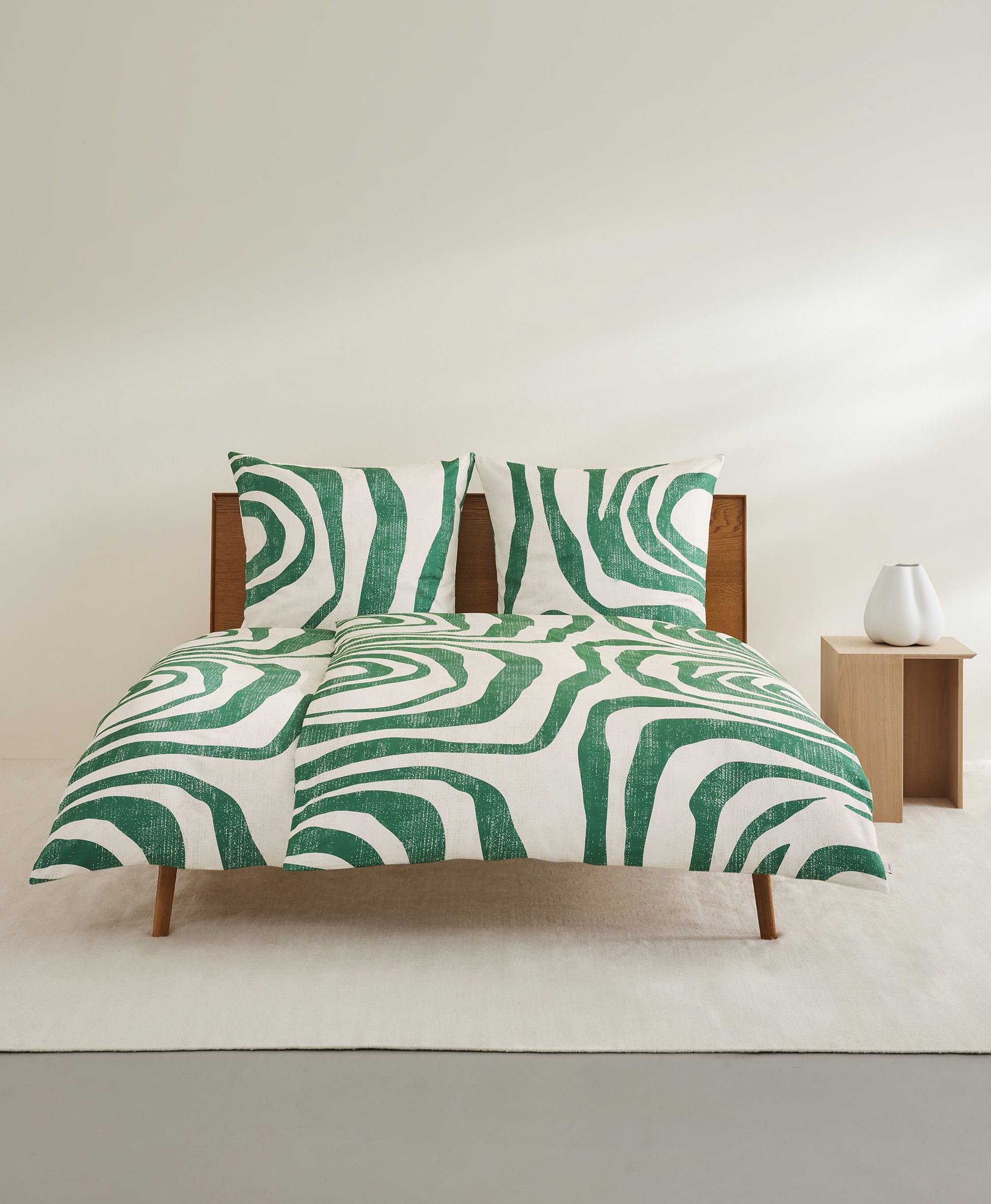 Auf einem Bett liegen mehrere Kopfkissen und eine Bettdecke bezogen mit Mako-Satin Bettwaesche aus 100 Prozent Baumwolle mit Muster aus grafischen Formen in der Farbe Gruen. Modell Green.