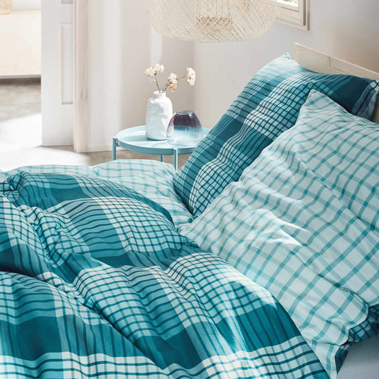 Auf einem Bett liegen mehrere Kopfkissen und eine Bettdecke bezogen mit Mako-Satin Bettwaesche aus 100 Prozent Baumwolle mit Muster aus Quadraten in der Farbe Tuerkis. Modell Karo.