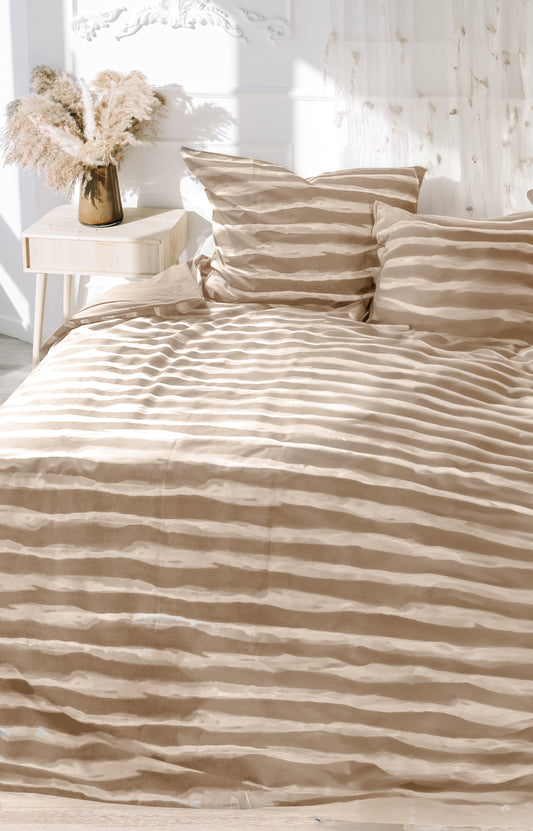 Auf einem Bett liegen mehrere Kopfkissen und eine Bettdecke bezogen mit Renforce Bettwaesche aus 100 Prozent Baumwolle mit Muster aus horizontalen Streifen in der Farbe Beige. Modell Sandra Stripes Beige.