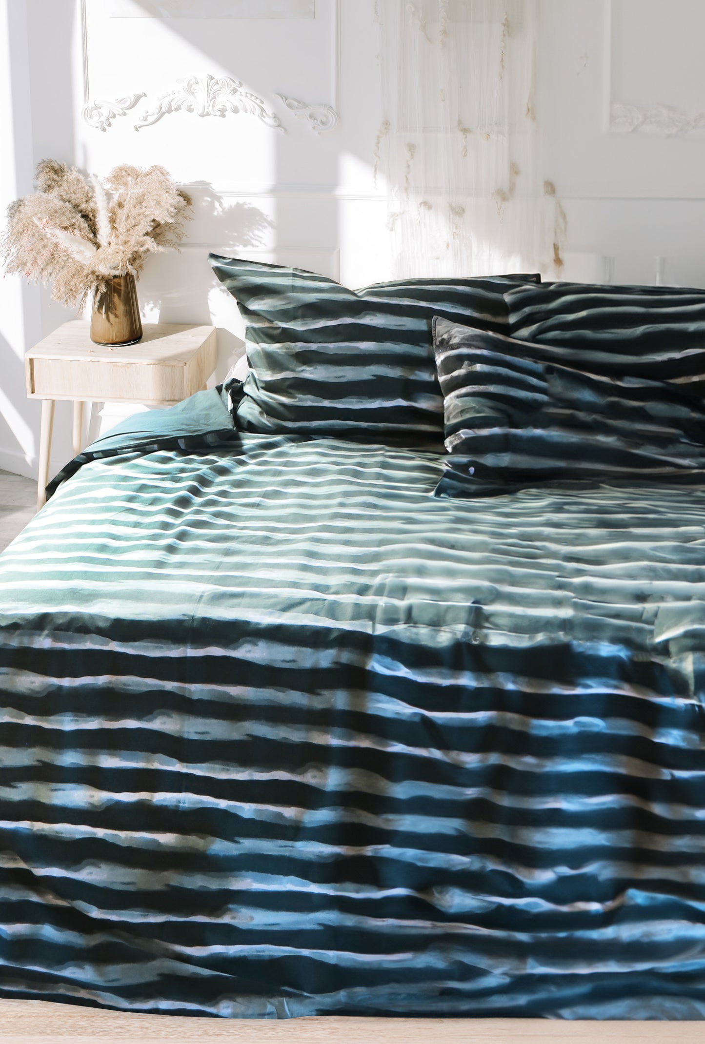 Auf einem Bett liegen mehrere Kopfkissen und eine Bettdecke bezogen mit Renforce Bettwaesche aus 100 Prozent Baumwolle mit Muster aus horizontalen Streifen in der Farbe Petrol. Modell Stripes Petrol.