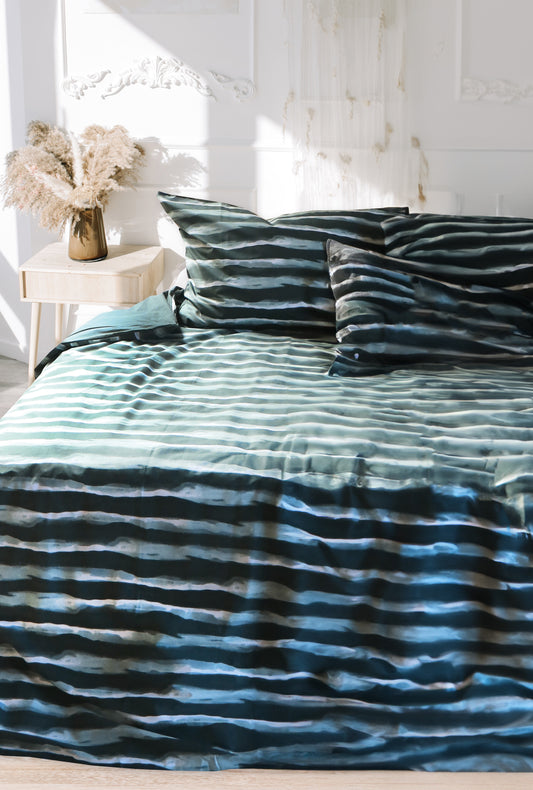 Auf einem Bett liegen mehrere Kopfkissen und eine Bettdecke bezogen mit Renforce Bettwaesche aus 100 Prozent Baumwolle mit Muster aus horizontalen Streifen in der Farbe Petrol. Modell Stripes Petrol.
