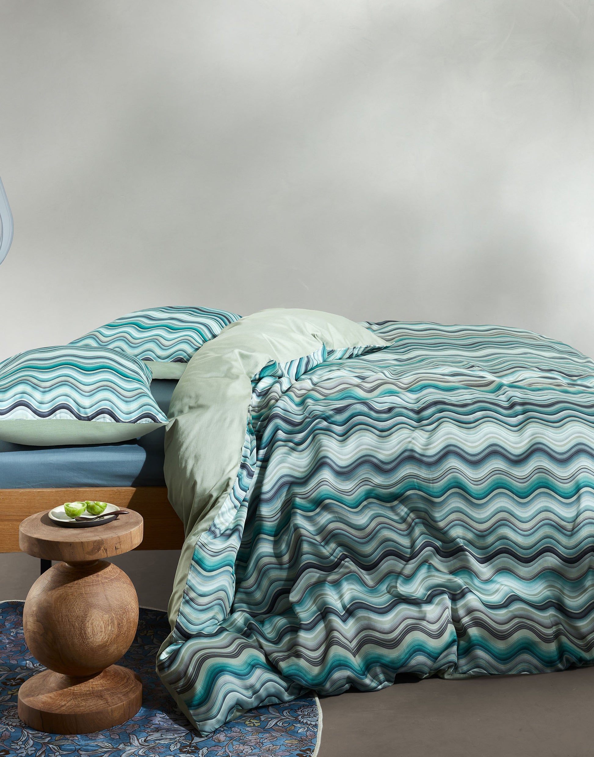Ein Bettbezug-Set aus mehreren Kopfkissen und einer Bettdecke bezogen mit Satin Bettwaesche aus 100 Prozent Baumwolle mit Muster aus vertikalen Schlangenlinien in der Farbe Tuerkis. Modell Sol Comforting Green.