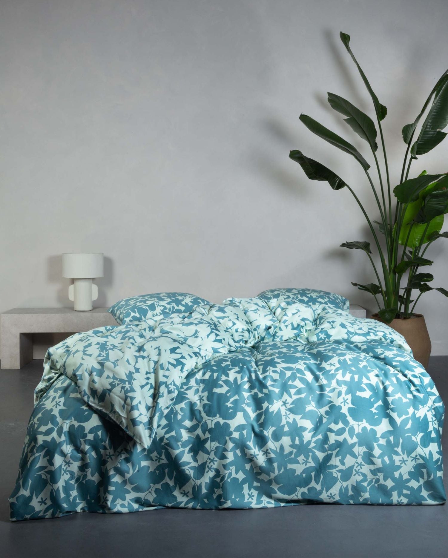 Ein Bettbezug-Set aus mehreren Kopfkissen und einer Bettdecke bezogen mit Satin Bettwaesche aus 100 Prozent Baumwolle mit Muster aus Blaettern in der Farbe Blau. Modell Sora Smoke Blue.