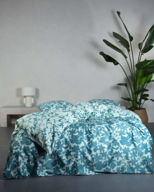 Ein Bettbezug-Set aus mehreren Kopfkissen und einer Bettdecke bezogen mit Satin Bettwaesche aus 100 Prozent Baumwolle mit Muster aus Blaettern in der Farbe Blau. Modell Sora Smoke Blue.