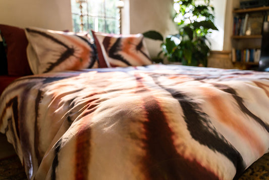 Auf einem Bett liegen mehrere Kopfkissen und eine Bettdecke bezogen mit Satin Bettwaesche aus 100 Prozent Baumwolle mit Muster aus grafischen Formen in der Farbe Rosegold. Modell Sidqi Russet.