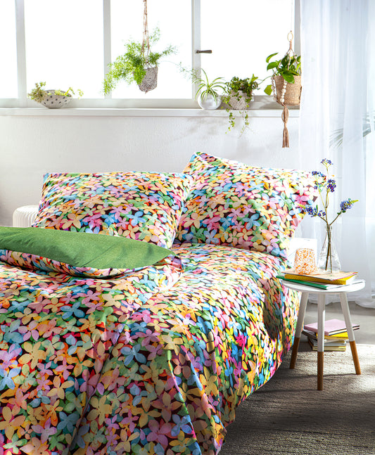 Auf einem Bett liegen mehrere Kopfkissen und eine Bettdecke bezogen mit Satin Bettwaesche aus 100 Prozent Baumwolle mit Muster aus Blumen in der Farbe Mehrfarbig. Modell Coloria.