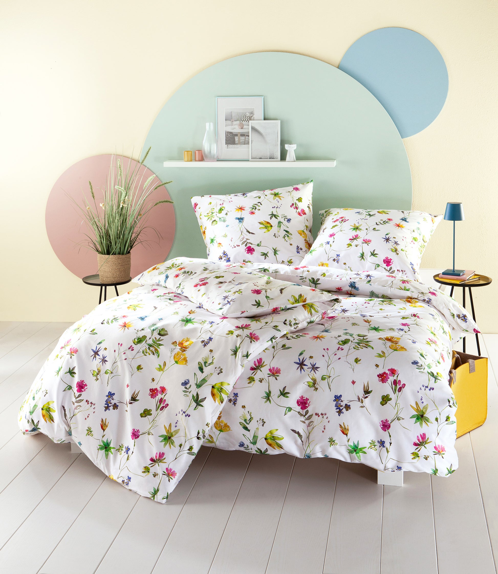 Auf einem Bett liegen mehrere Kopfkissen und eine Bettdecke bezogen mit Satin Bettwaesche aus 100 Prozent Baumwolle mit Muster aus Blumen in der Farbe Weiss. Modell Floravie.
