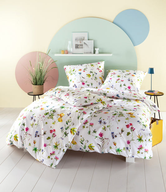 Auf einem Bett liegen mehrere Kopfkissen und eine Bettdecke bezogen mit Satin Bettwaesche aus 100 Prozent Baumwolle mit Muster aus Blumen in der Farbe Weiss. Modell Floravie.
