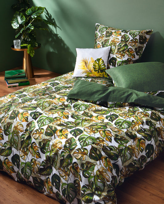 Auf einem Bett liegen mehrere Kopfkissen und eine Bettdecke bezogen mit Satin Bettwaesche aus 100 Prozent Baumwolle mit Muster aus Blaettern in der Farbe Gruen. Modell Verdea.