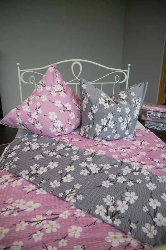 Auf einem Bett liegen mehrere Kopfkissen und eine Bettdecke bezogen mit Satin-Seersucker Bettwaesche aus 100 Prozent Baumwolle mit Muster aus Blueten in der Farbe Grau. Modell Grau.