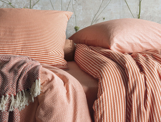 Auf einem Bett liegen mehrere Kopfkissen und eine Bettdecke bezogen mit Soft Seersucker Bettwaesche aus 100 Prozent Baumwolle mit Muster aus vertikalen Linien in der Farbe Koralle. Modell Shadow Orange.
