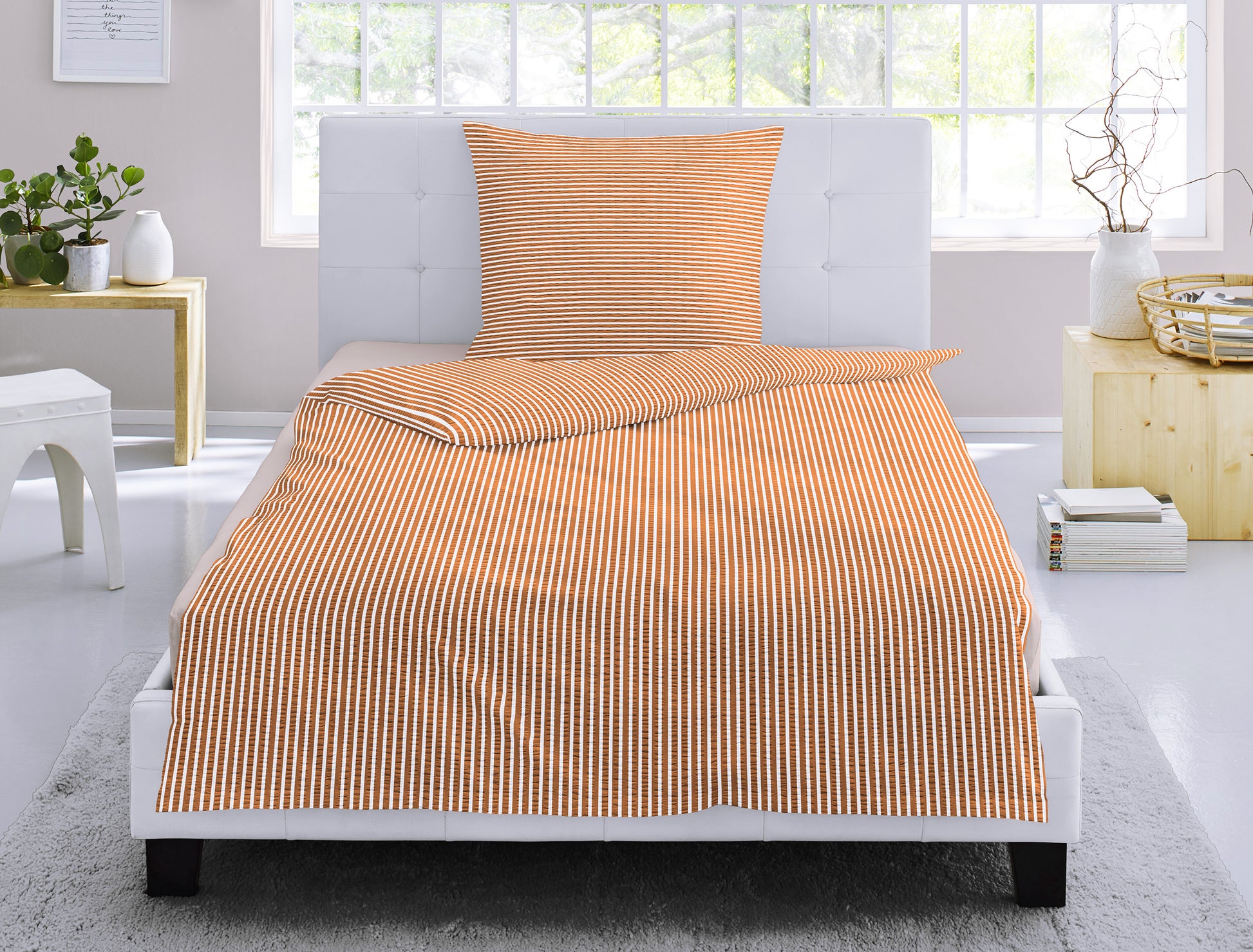 Auf einem Bett liegt ein Kopfkissen und eine Bettdecke bezogen mit Soft Seersucker Bettwaesche aus 100 Prozent Baumwolle mit Muster aus vertikalen Linien in der Farbe Koralle. Modell Shadow Orange Stripe.