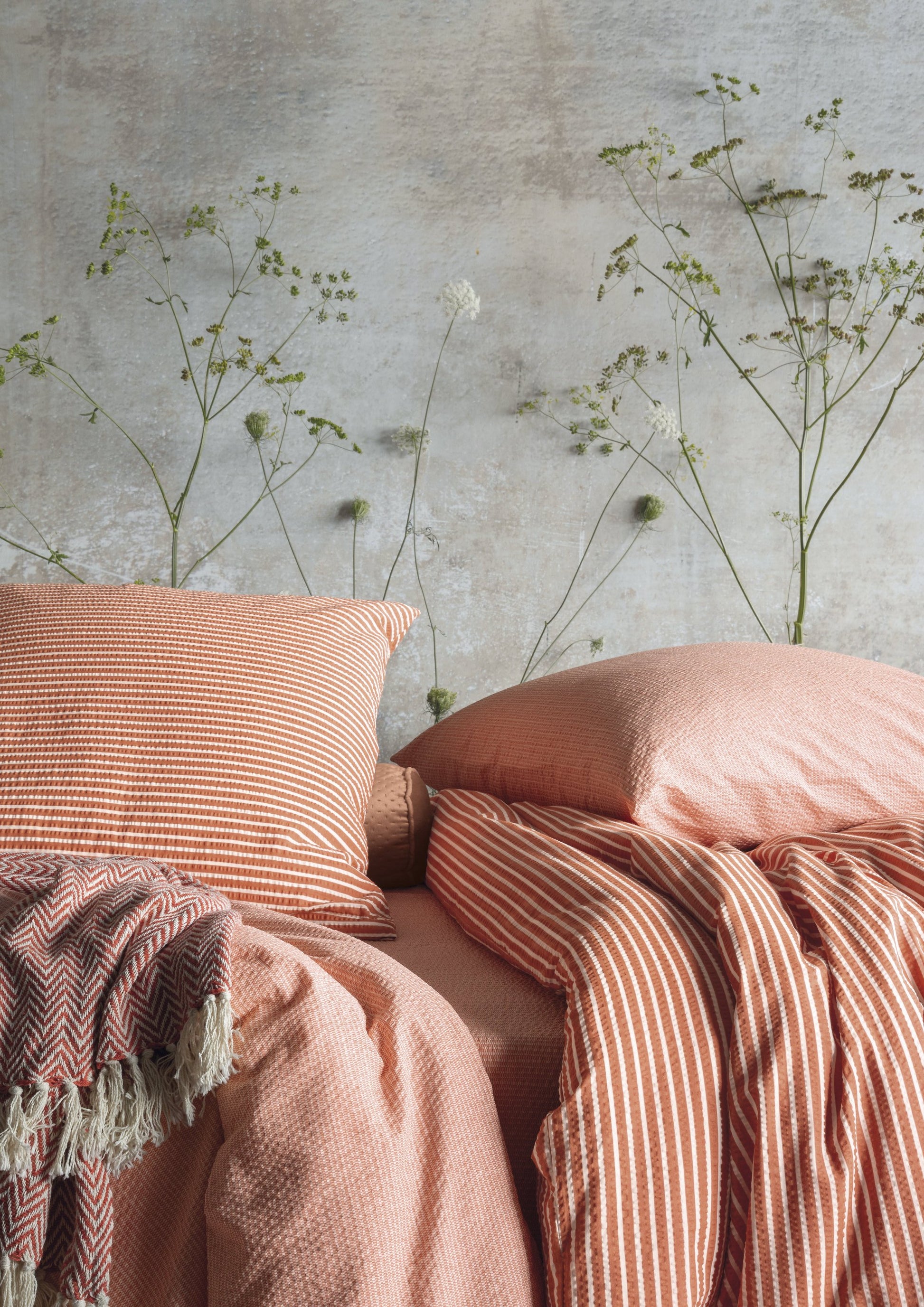Auf einem Bett liegen mehrere Kopfkissen und eine Bettdecke bezogen mit Soft Seersucker Bettwaesche aus 100 Prozent Baumwolle mit Muster aus vertikalen Linien in der Farbe Koralle. Modell Shadow Orange Stripe.