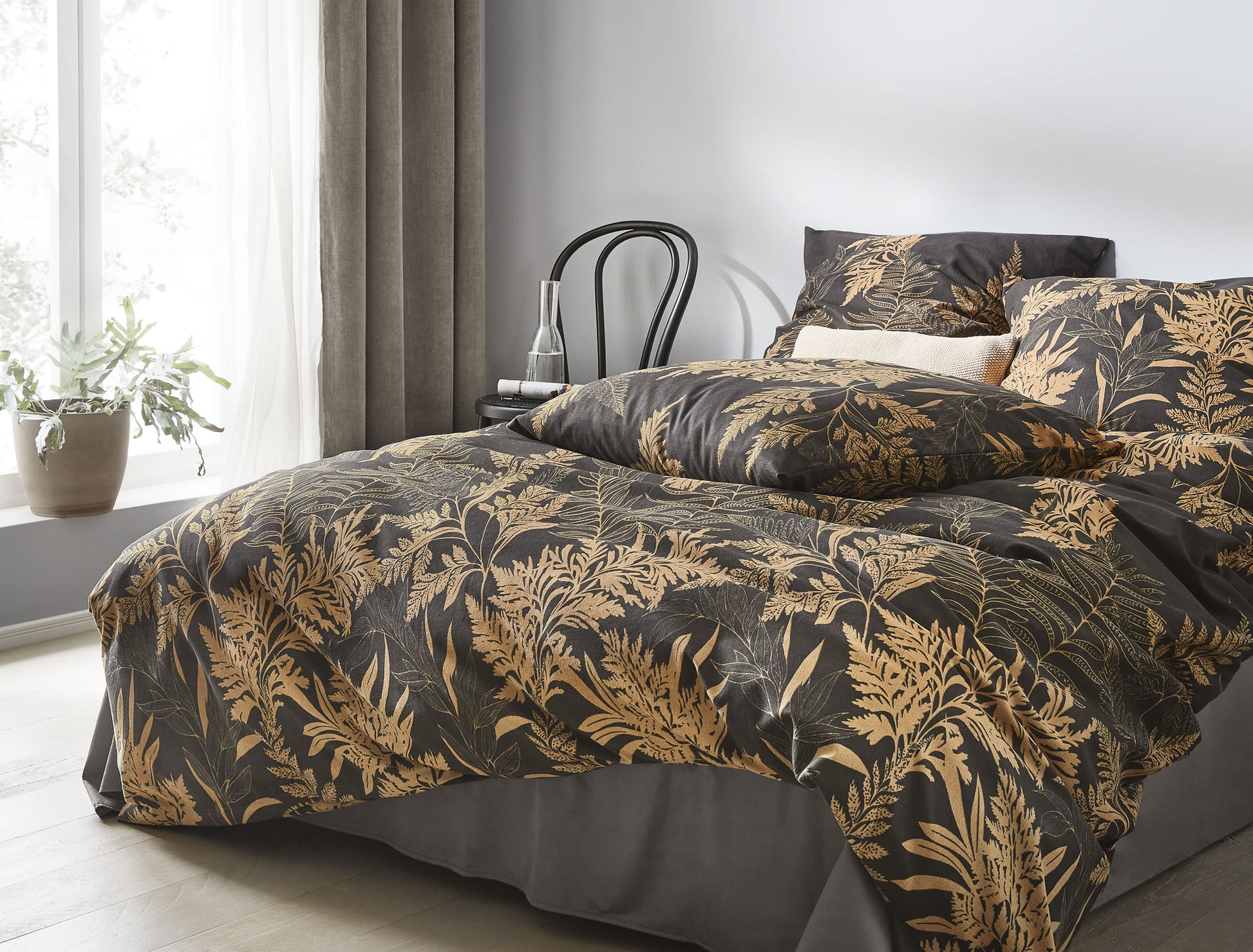 Auf einem Bett liegen mehrere Kopfkissen und eine Bettdecke bezogen mit Edel-Feinbiber Bettwaesche aus 100 Prozent Baumwolle mit Muster aus Farnen in der Farbe Braun. Modell Farn.